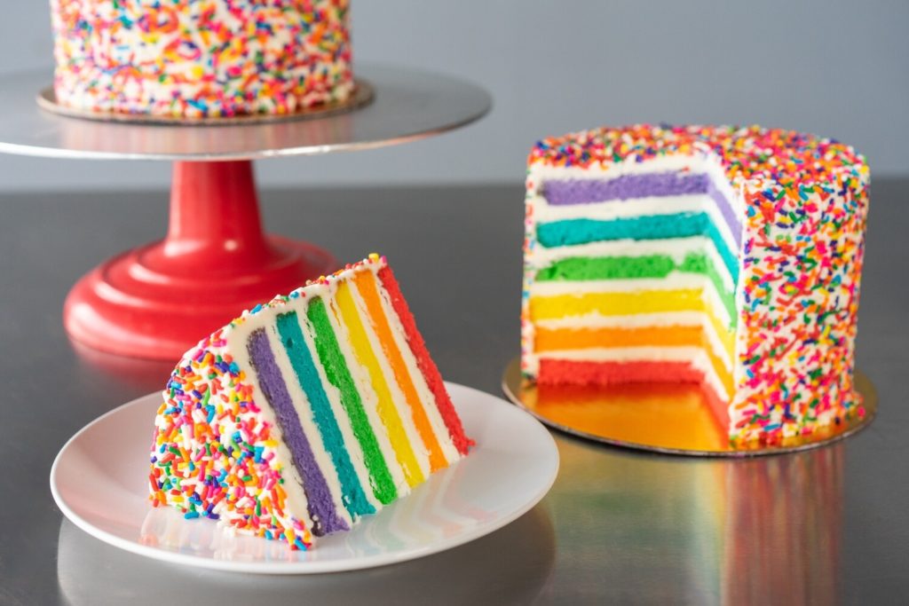 Image of a rainbow cake with a slice cut out from Carlo's Bakery.