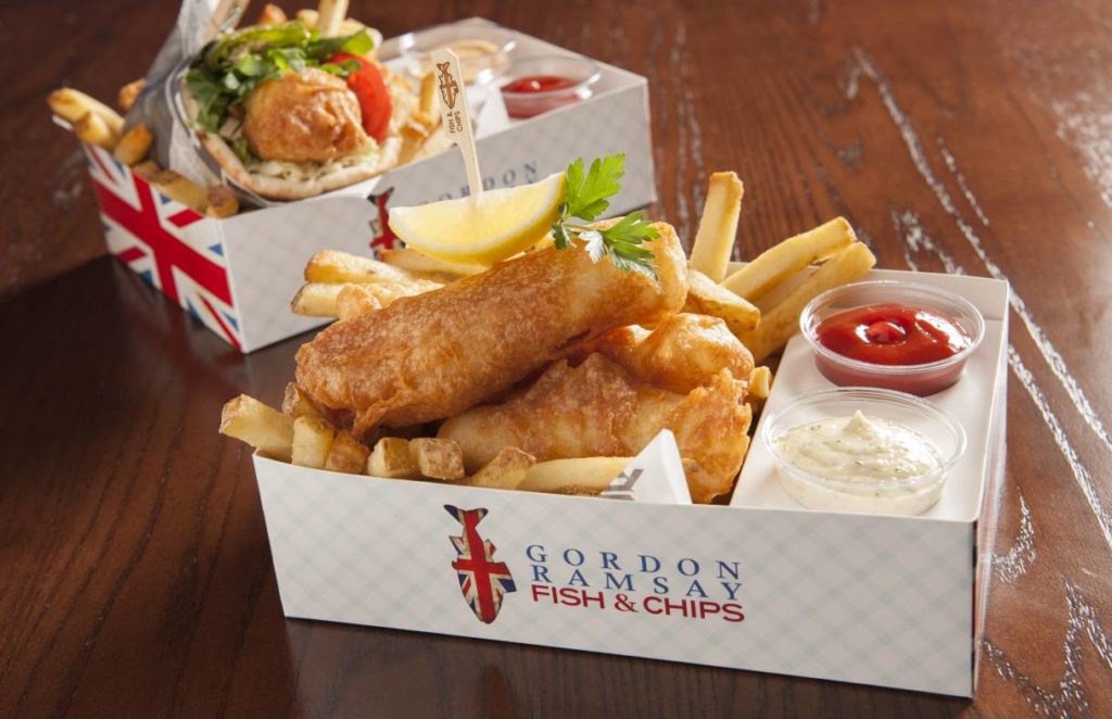 gordon ramsay's fish n chips on display over a bed of fries and sides of condiments 