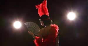 a burlesque dancer with a red feather boa and headress waving a fan on a dark stage with spotlights