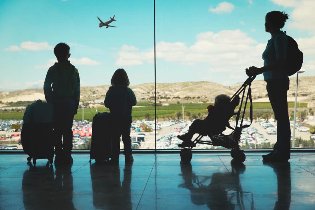 a mother with a stroller and baby looking out the airport window, with two kids and their suitcases
