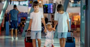 Three children holding hands while walking through an airport; two older children on either side of a toddler help it walk.