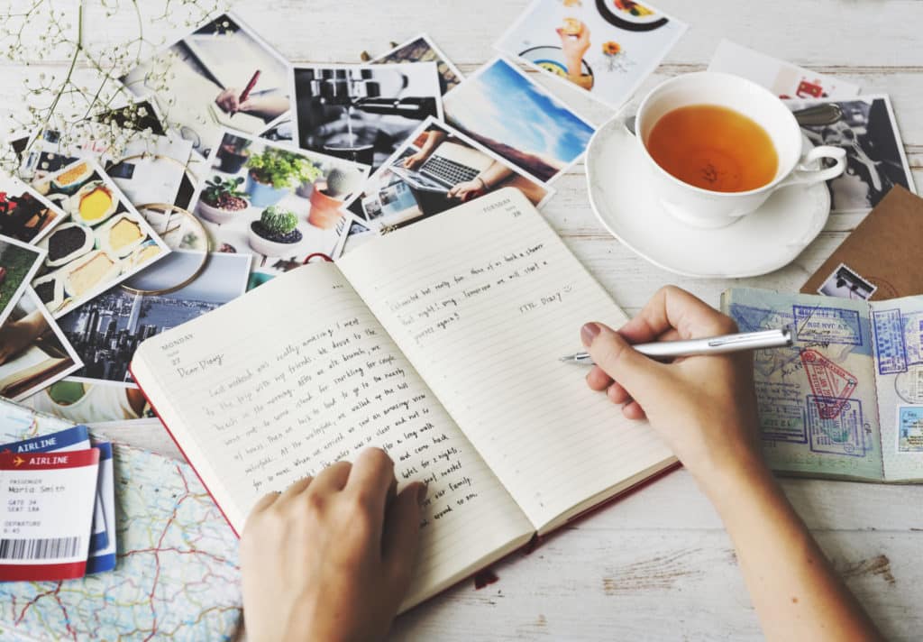 hands writing in a journal with a cup of tea and airline tickets among pictures