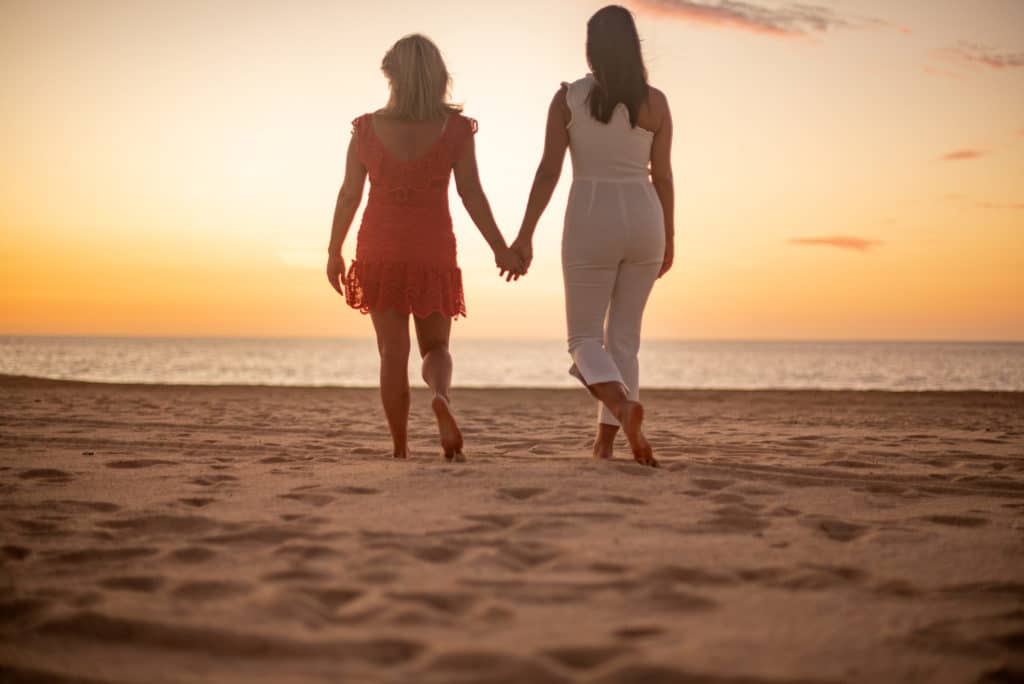 two women holding hands and walking on a Cabo beach at sunset